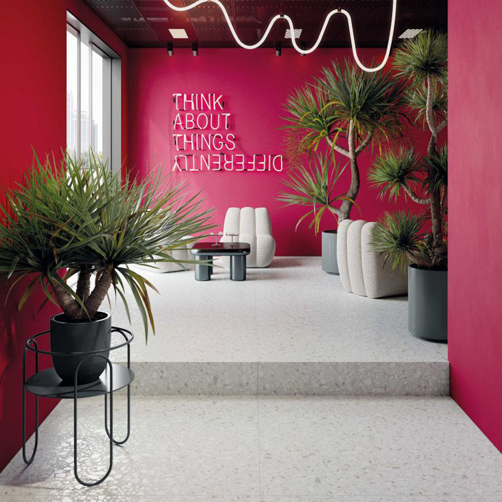 Commercial interior featuring Shards Blanco 3D Terrazzo 60x120cm porcelain floor tiles, showcasing a white terrazzo effect with textured 3D stone chips, paired with bold pink walls, contemporary seating, statement lighting and tropical plants for a striking modern space.