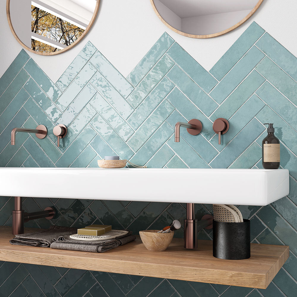 Nomad Aquamarine brick wall tiles in a 6 × 24.6cm format, laid in a herringbone pattern with a glossy, lightly textured finish, shown in a bathroom setting with soft blue-green tones and an industrial-style look.