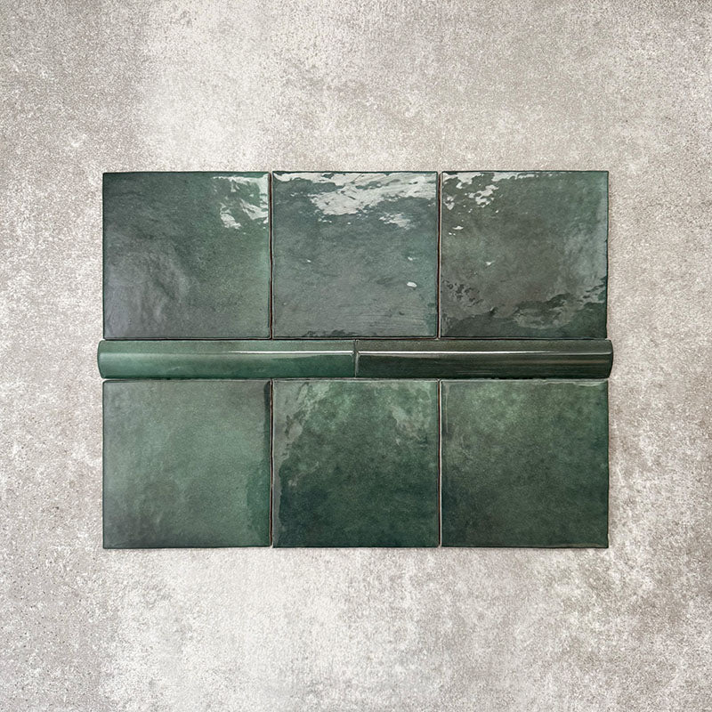 Flat lay showing Zellige Royal Green Square 13.2 × 13.2cm Moroccan-style ceramic tiles paired with a matching Royal Green Bullnose 3 × 20cm trim tile in rich deep green tones, arranged on a textured grey background to highlight the glossy glaze, subtle tonal variation and handcrafted zellige-effect finish.