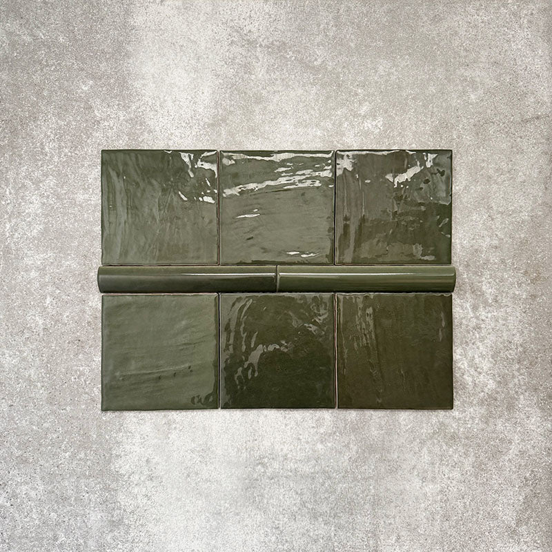Flat lay featuring Zellige Botanical Green Square tiles with matching bullnose trim, showcasing 13.2 × 13.2cm Moroccan-style ceramic squares and a 3 × 20cm rounded bullnose edge in deep botanical green tones, with a glossy glaze, subtle tonal variation and handcrafted zellige-effect finish on a neutral textured background.