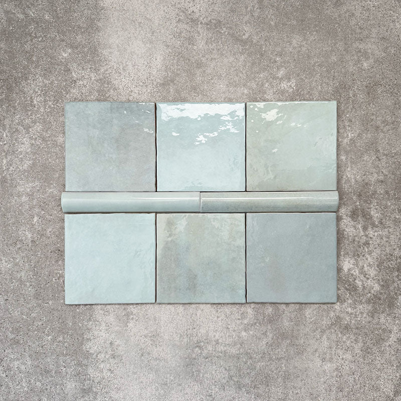Flat lay of Zellige Aqua Moroccan-style ceramic tiles, showing 13.2×13.2cm square tiles paired with a 3×20cm bullnose trim, highlighting the rounded edge detail, glossy blue-green glaze and natural tonal variation on a neutral background.