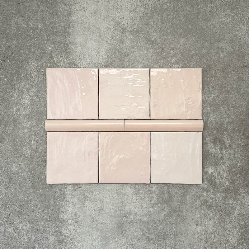 Flat lay showing Zellige Pink Mist Square 13.2 × 13.2cm Moroccan-style ceramic tiles paired with a matching Pink Mist Bullnose 3 × 20cm trim tile in soft pale pink tones, arranged on a textured grey background to highlight the glossy glaze, subtle tonal variation and handcrafted zellige-effect finish.