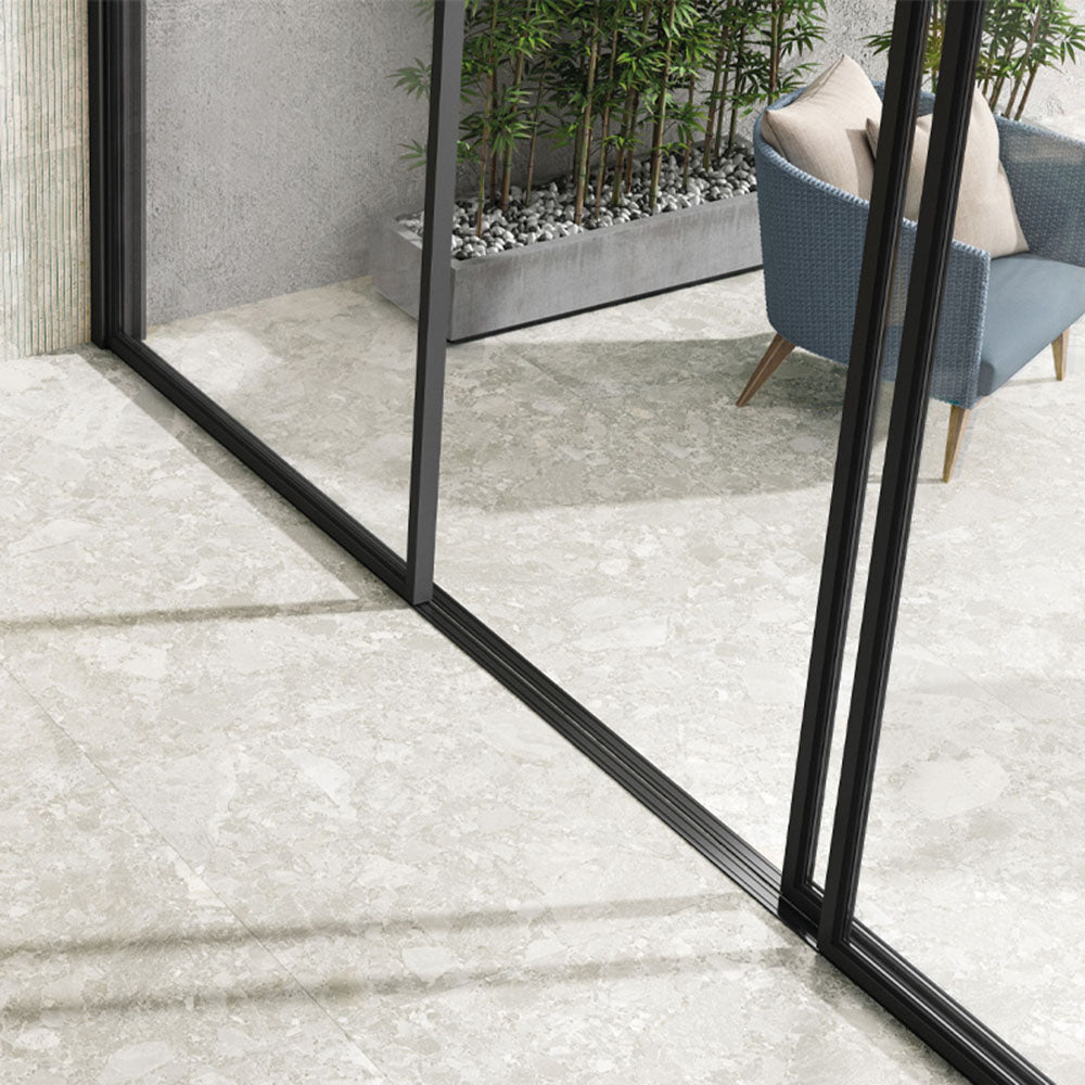 Indoor-outdoor space featuring Essentials Terrazzo Stone Blanca 60x120cm terrazzo effect porcelain floor tiles, paired with sliding glass doors, a woven lounge chair, and planted greenery for a seamless, light-filled finish.