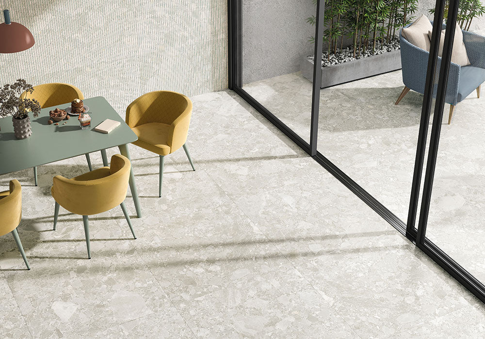 Dining area featuring Essentials Terrazzo Stone Blanca 60x120cm terrazzo effect porcelain floor tiles, paired with a green table, mustard chairs, textured wall tiles, and large sliding glass doors for a light, open feel.