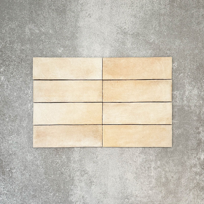 Flat lay of Zellige Arena Marfil Brick 6.5 × 20cm Moroccan-style ceramic tiles, shown in a warm beige and cream colour palette with subtle tonal variation and gently uneven edges, arranged in a vertical brick layout on a textured grey background to highlight the handcrafted, zellige-effect finish.