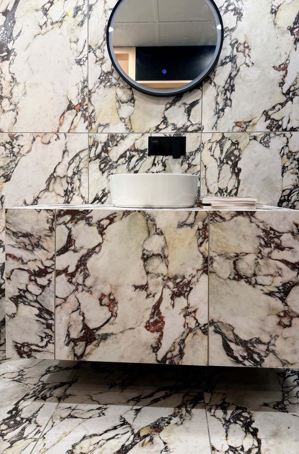 Bathroom vanity area featuring Viola Matt Honed Marble effect 60 × 60cm porcelain wall and floor tiles, styled with a floating basin, black wall-mounted tap, and bold veining in cream, burgundy, and charcoal tones.