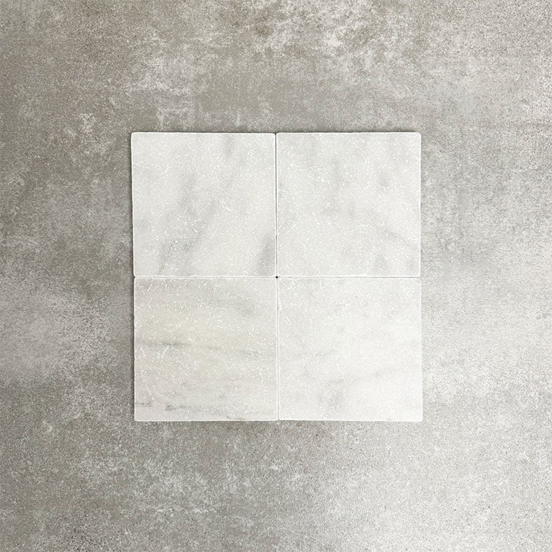 Four Fayrouz White 15.2x15.2cm natural marble tiles with a tumbled finish arranged together, showcasing soft white tones, subtle veining and natural variation against a neutral textured background.