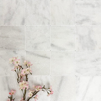 Flat lay of Fayrouz White 15.2x15.2cm natural marble tiles with a tumbled finish, featuring soft white tones and subtle natural veining, styled with delicate pink blossom branches for a calm, elegant look.