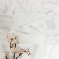 Flat lay of Fayrouz White 15.2x15.2cm natural marble tiles with a tumbled finish, featuring soft white tones and subtle natural veining, styled with delicate pink blossom branches for a calm, elegant look.