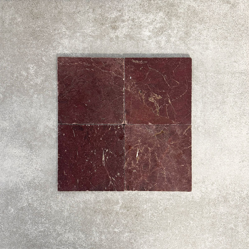 Four Fayrouz Burgundy Red 15.2x15.2cm natural marble tiles with a tumbled finish arranged together, showcasing rich red tones, natural veining and surface variation against a neutral textured background.