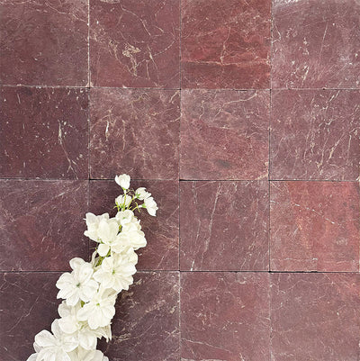 Flat lay of Fayrouz Burgundy Red 15.2x15.2cm natural marble tiles with a tumbled finish, showing rich red tones and natural veining, styled with soft white flowers for contrast and texture.
