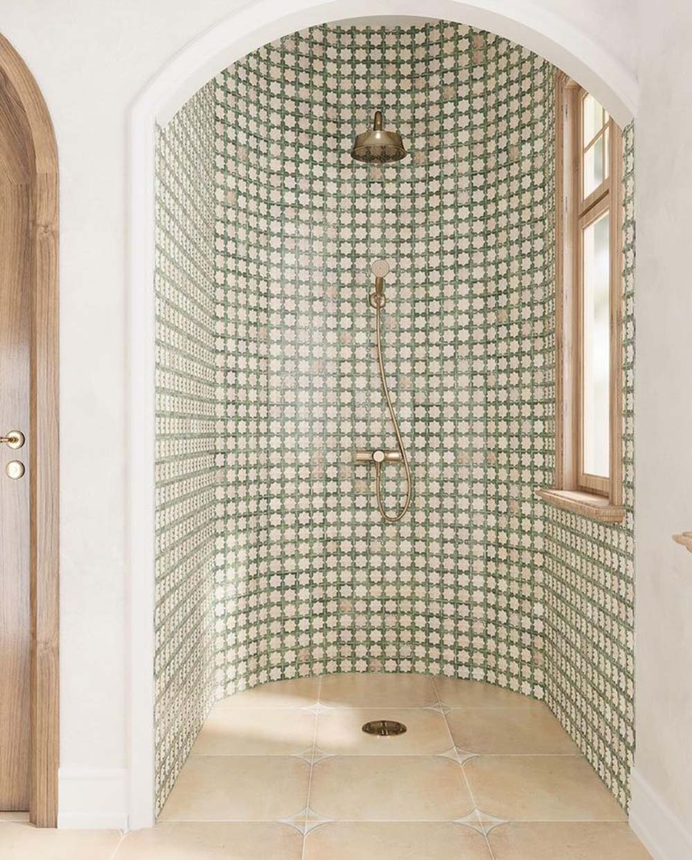 Walk-in shower featuring Zanzibar Moroccan Emerald Green ceramic wall tiles in a repeating Moroccan style star pattern, paired with warm stone effect floor tiles, brushed brass shower fittings, an arched alcove and natural wood window frames for a bright Mediterranean-inspired bathroom.