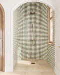 Walk-in shower featuring Zanzibar Moroccan Emerald Green ceramic wall tiles in a repeating Moroccan style star pattern, paired with warm stone effect floor tiles, brushed brass shower fittings, an arched alcove and natural wood window frames for a bright Mediterranean-inspired bathroom.